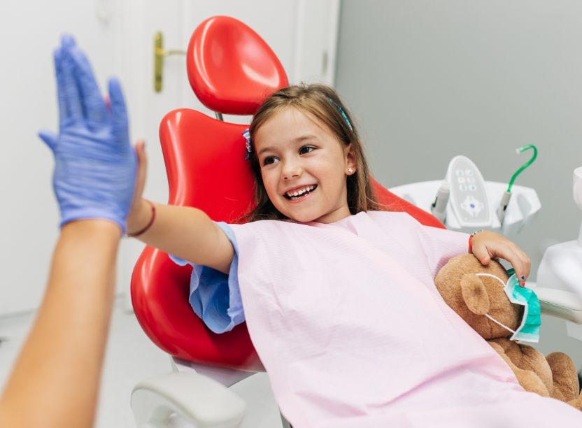 Little girl with teddy bear in dentist's chair, high-fiving dentist